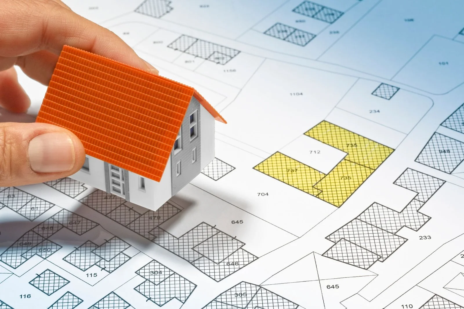 Our Services 4 Hand placing a miniature house model on an illustrated cadastral map with marked property boundaries, symbolizing land surveying, real estate, or plot selection.